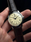 Montre Bambino tenue à la main, cadran crème et bracelet cuir marron
